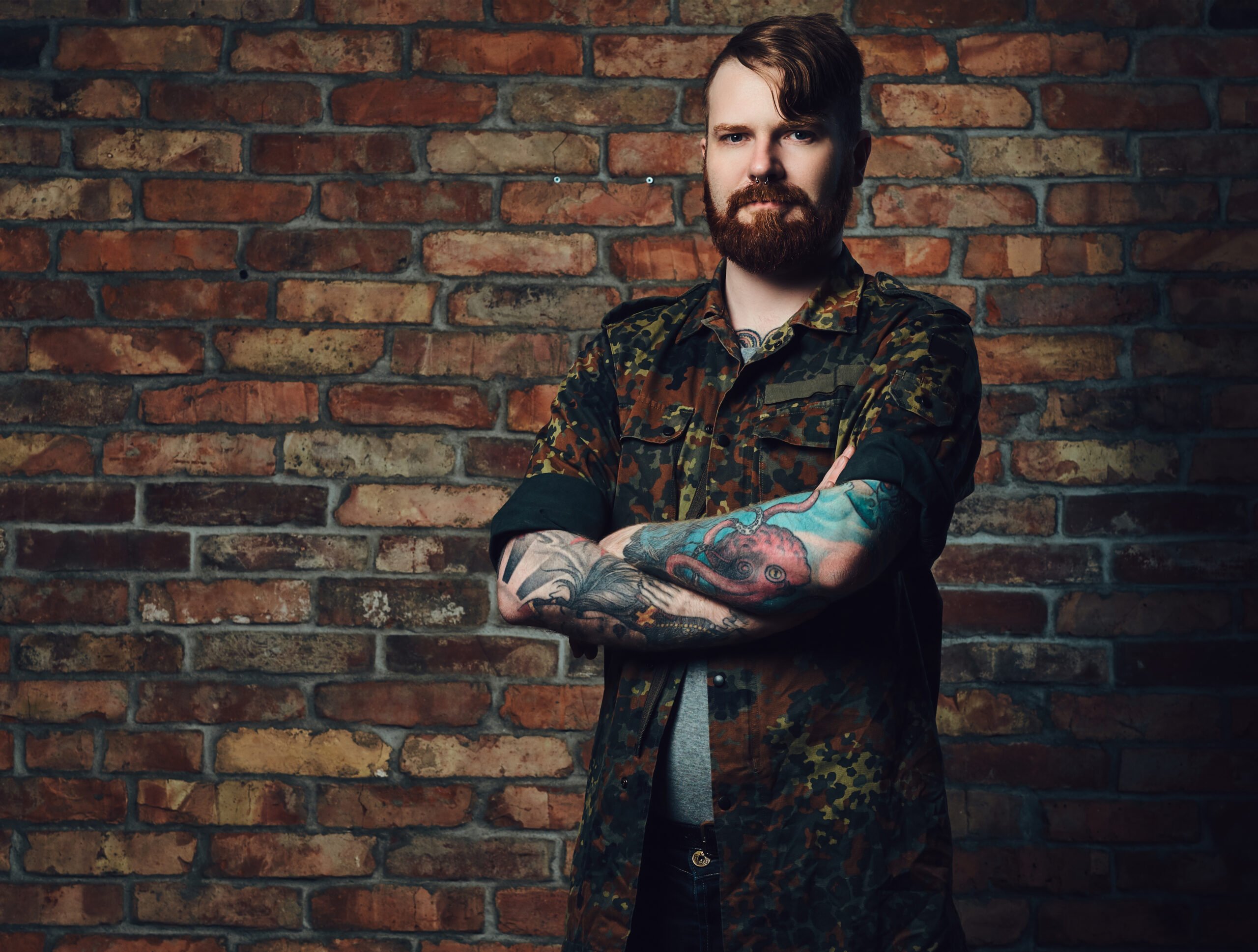 portrait-redhead-bearded-male-with-tattoos-arms-dressed-military-jacket-wall-brick-scaled.jpg